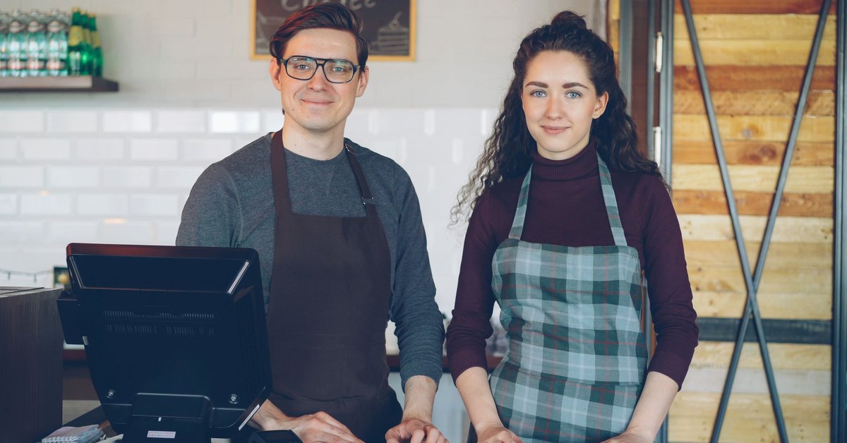 Two store employees in aprons standing behind a service counter next to a point-of-sale terminal